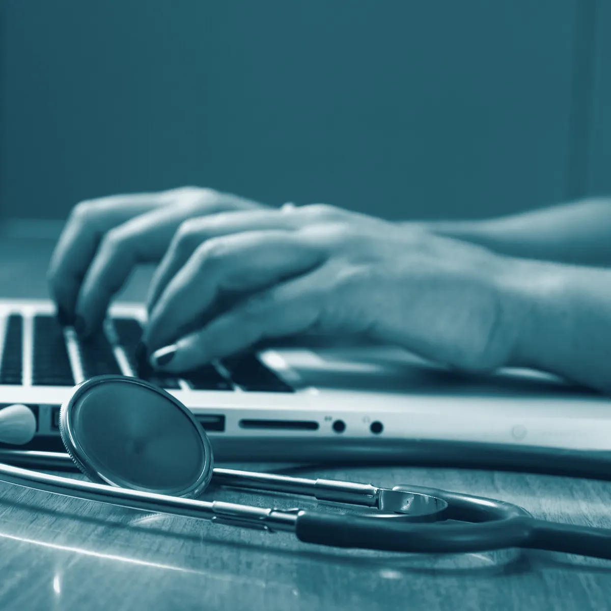 Mãos digitando no computador do consultório, com estetoscópio ao lado